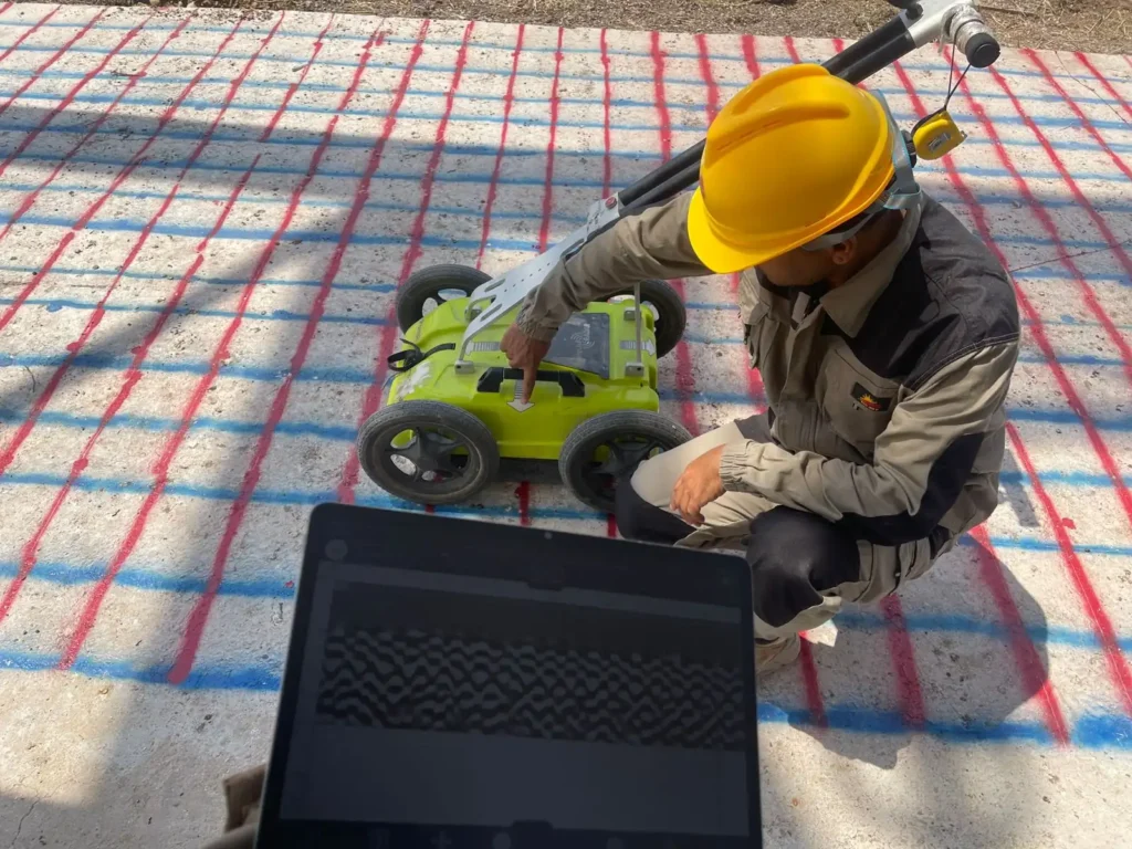Rebar mesh marked out in red and blue across the concrete slab, with the live radargram on the laptop beside the GPR cart